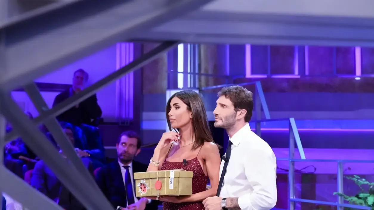 Due persone su un set televisivo di Affari Tuoi, una tiene in mano un pacco dorato