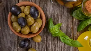 Ciotola di olive verdi e nere accanto a foglie di basilico e bottiglia di olio su tavolo rustico