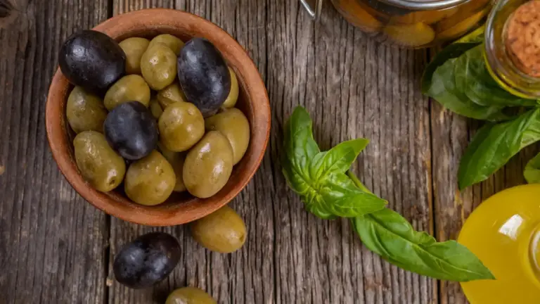 Ciotola di olive verdi e nere accanto a foglie di basilico e bottiglia di olio su tavolo rustico